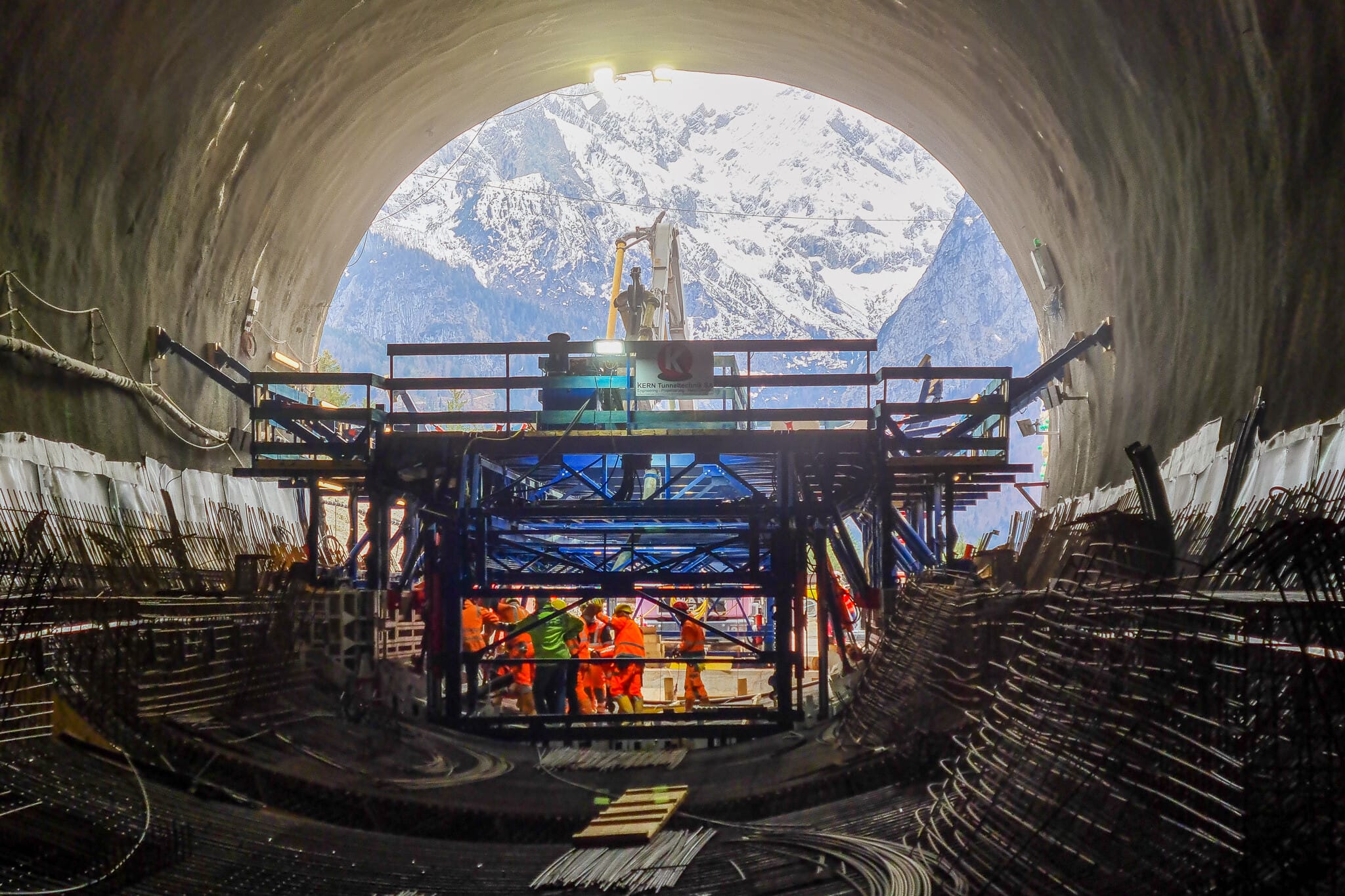 Blick aus den Ausgang einer im Bau befindlichen Tunnelröhre auf die dahinterliegenden Berge mit Bauarbeitern und Stahlbewehrung im Vordergrund