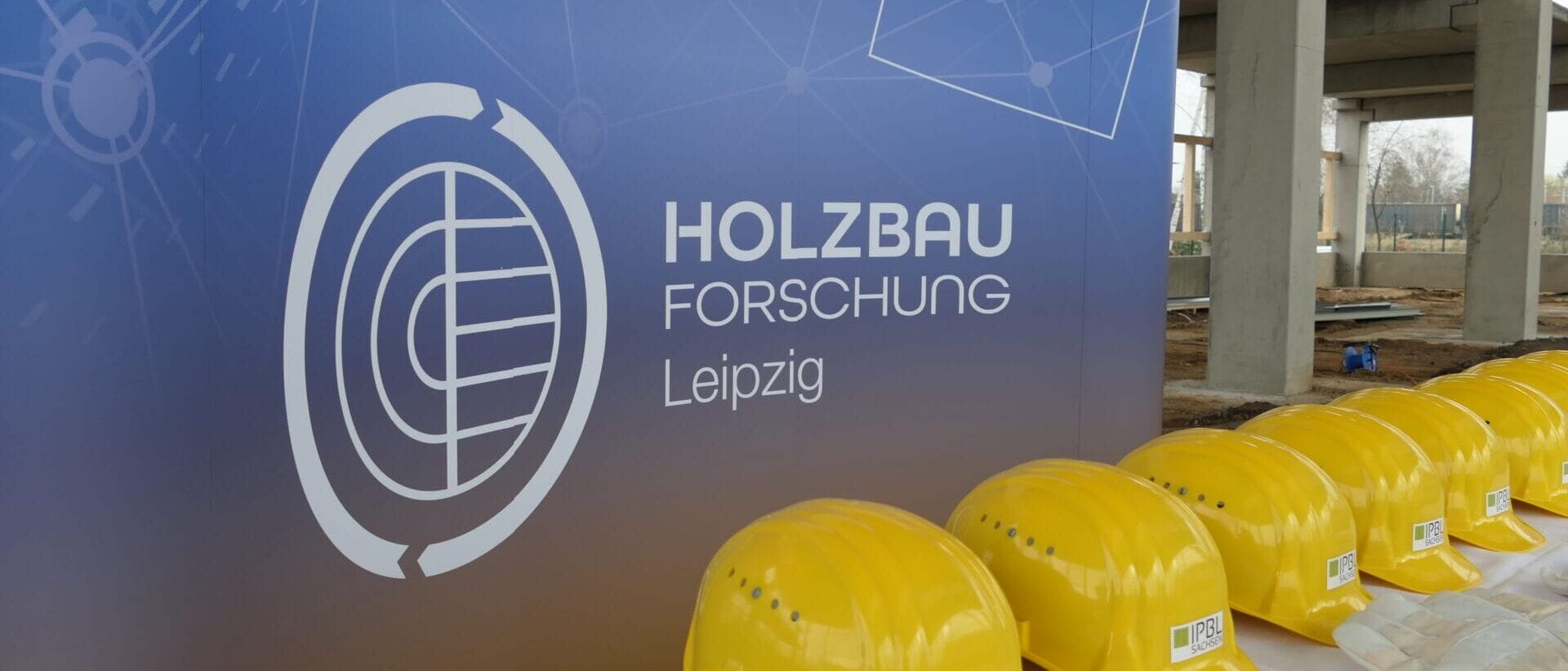 gelbe Bauhelme des InnovationsParks Bautechnik Leipzig Sachsen vor dem weißen Logo der HOLZBAU Forschung Leipzig